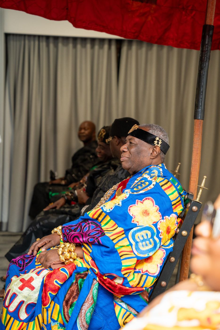A chief in kente and gold regalia seated beneath a red state umbrella