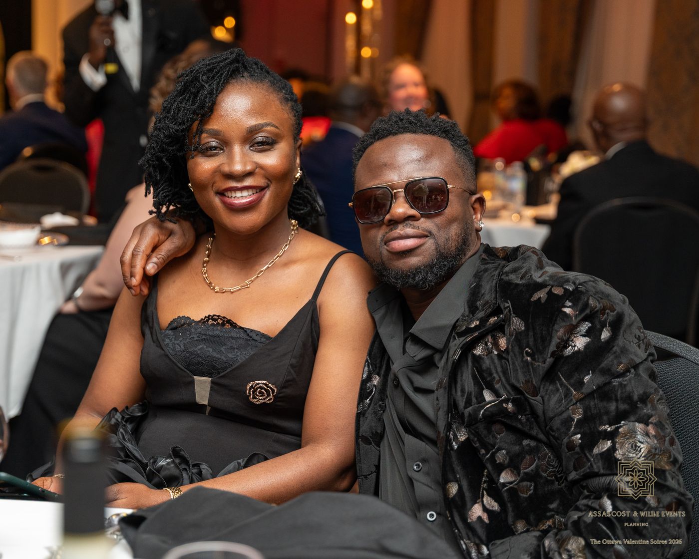 A couple smiling at their table, one in sunglasses and a velvet jacket