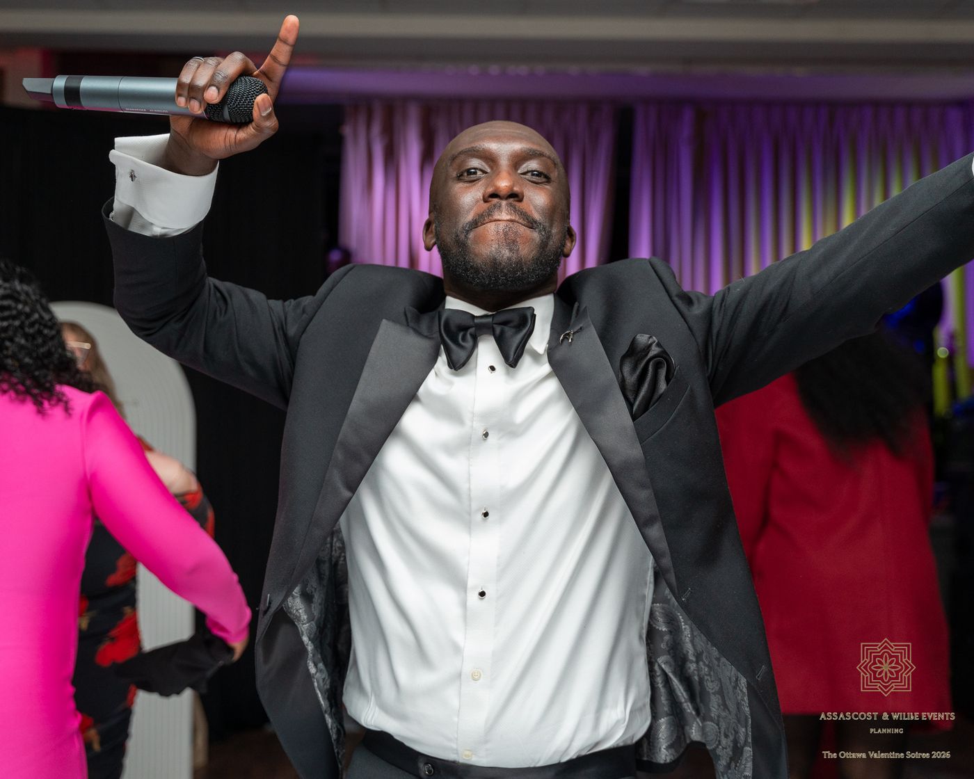 The host in a tuxedo with his arms raised, holding a microphone