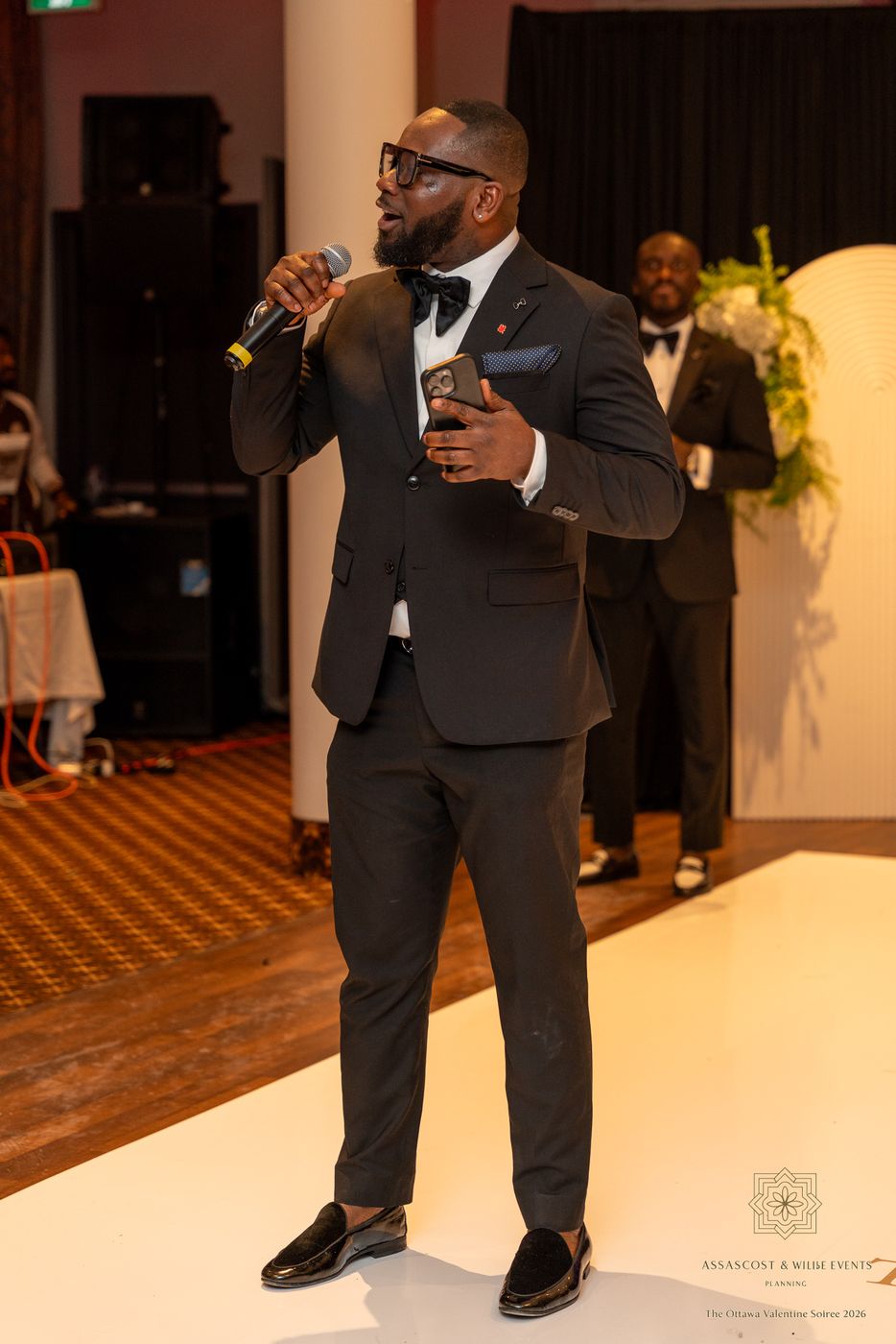 A host in glasses and a bow tie speaking into the microphone