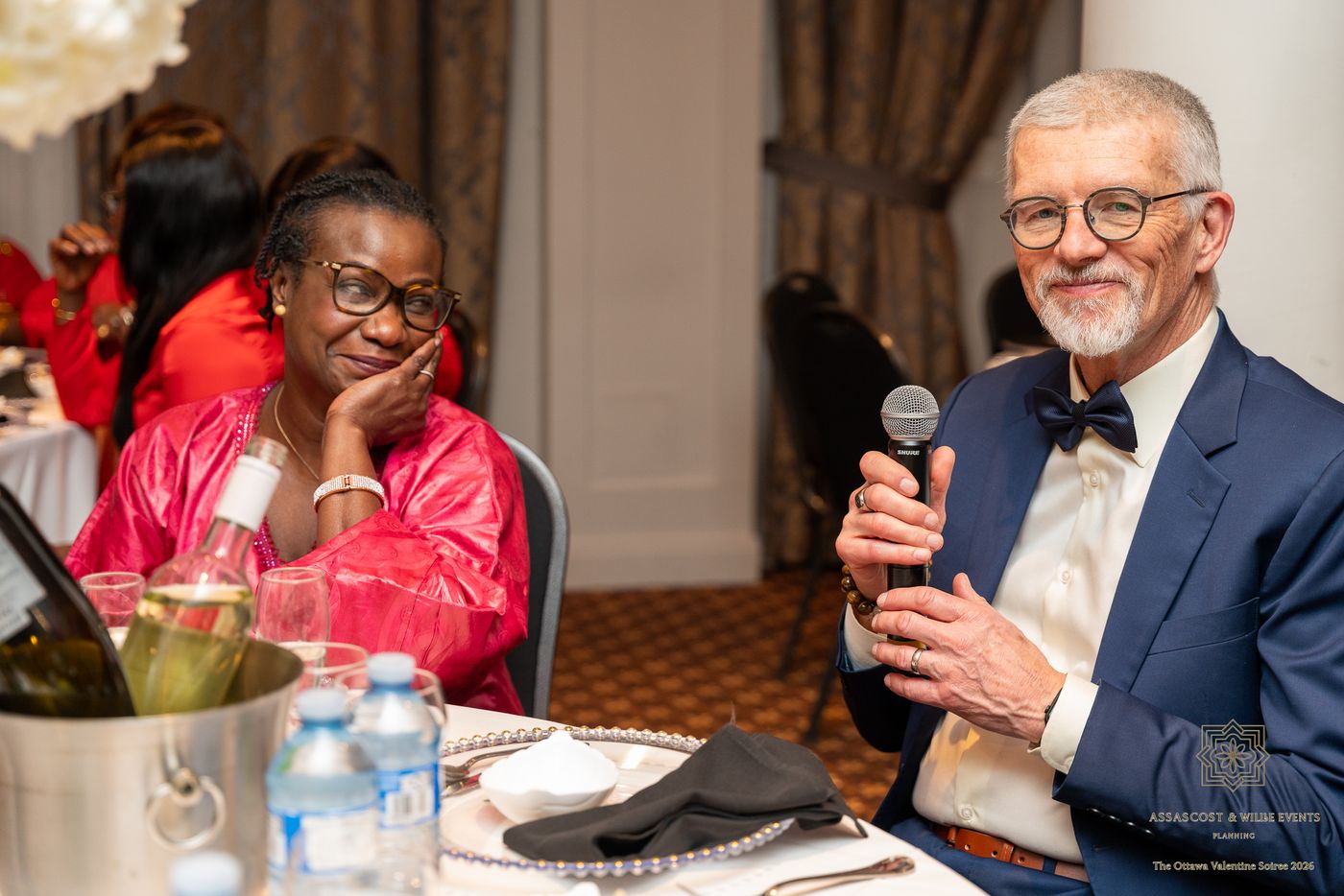 A guest in a navy suit and bow tie holding a microphone beside a smiling guest in pink